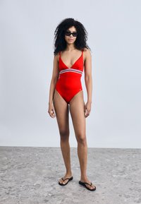 Κόκκινο ολόσωμο μαγιό με βαθύ V λαιμόκοψη, λευκές και μαύρες οριζόντιες ρίγες στη μέση, συνοδευόμενο από μαύρα σαγιονάρες.