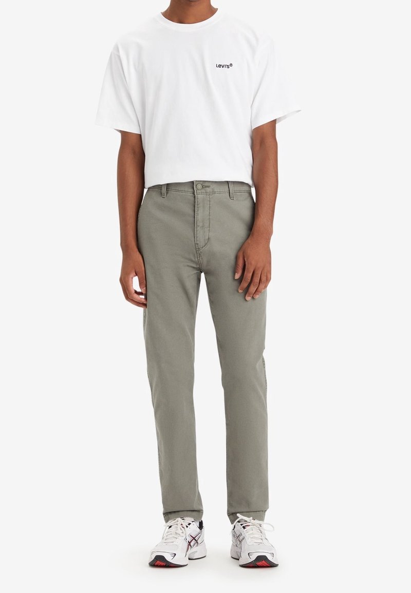 Biała koszulka z krótkim rękawem z czarnym logo „Levi's”, zestawiona z jasnooliwkowymi spodniami i białymi tenisówkami z czerwonymi akcentami.