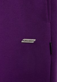 Lila Stoff mit gerippter Textur, ausgestattet mit einem silbernen Etikett, das mit "SMILODOX" geprägt ist, sowie einem schwarzen Seitenetikett.