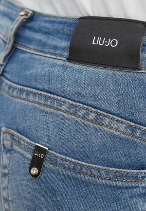 Jeans di denim blu con una toppa in pelle nera e una targhetta metallica, entrambe recanti il nome del marchio "LIU JO" sulla parte posteriore della cintura e sulla tasca.