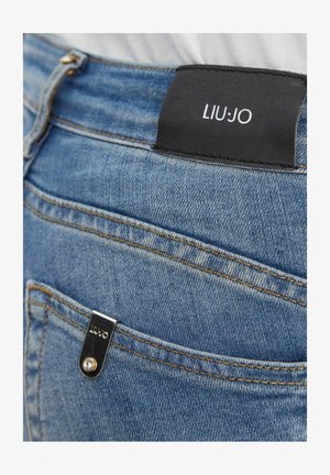 Jeans di denim blu con una toppa in pelle nera e una targhetta metallica, entrambe recanti il nome del marchio "LIU JO" sulla parte posteriore della cintura e sulla tasca.