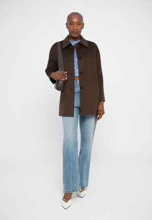 Cappotto di lana marrone con colletto, chiusura a bottoni e tasche laterali; indossato sopra una camicia blu e jeans chiari svasati con scarpe bianche a punta.