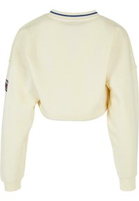 Cropped sweatshirt in cream, featuring long sleeves, a ribbed collar with blue accent, and a logo patch on the left sleeve.