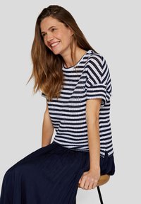 Navy and white striped knit t-shirt with short sleeves and round neckline, worn with a pleated navy skirt, seated on a stool.