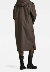 Brązowy, długi płaszcz z kapturem o lekkim połysku, z bocznym rozporkiem i naszywką z materiału. Do tego czarne, sięgające kolan buty.
