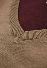 Beige Strickpullover mit V-Ausschnitt, der ein weinrotes Innenfutter aufweist. Der Stoff hat eine weiche Textur und gerippte Kanten für einen Kontrast.