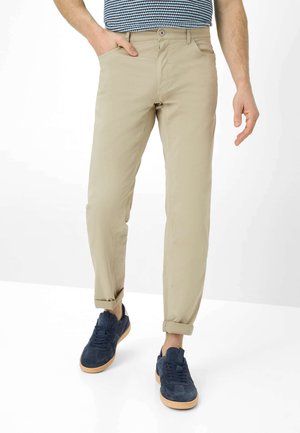 Homme portant un pantalon beige retroussé, une chemise rayée bleu et blanc rentrée, et des baskets bleu marine avec des semelles marron clair.