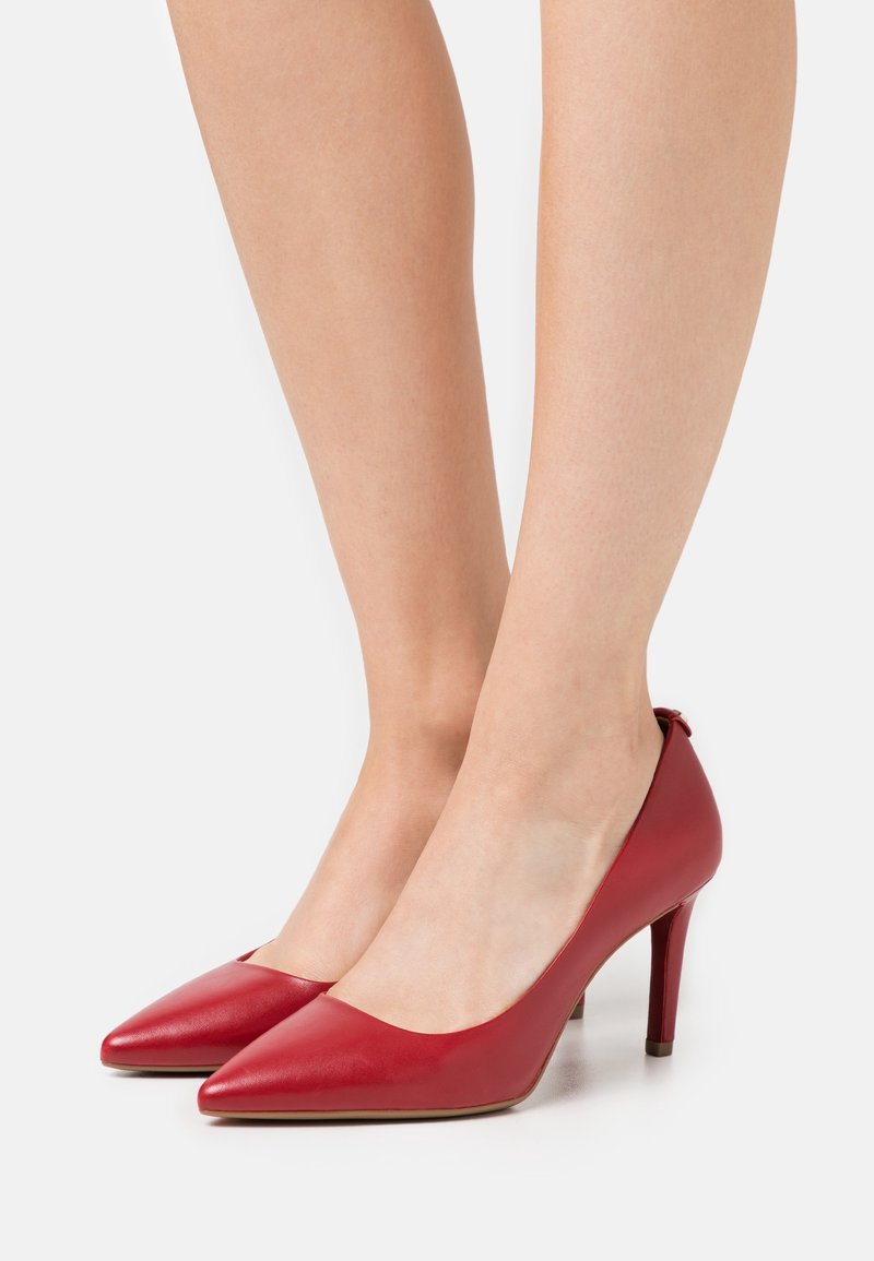 Paire de jambes portant des escarpins rouges à bout pointu avec des talons aiguilles fins sur un fond clair uni.