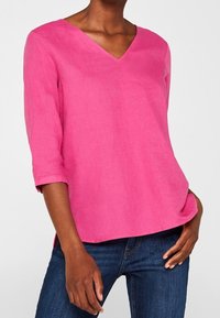 Blusa di lino rosa con scollo a V, maniche a tre quarti e taglio rilassato. Il tessuto appare leggero e ha una texture liscia.