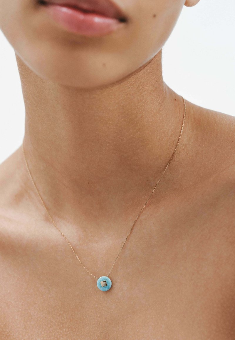 Collar de cadena de oro con un colgante circular de turquesa y un pequeño detalle cuadrado. Textura suave, diseño minimalista.