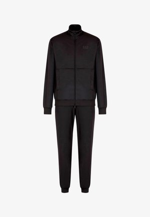 Chándal negro hecho de tejido suave. Presenta una chaqueta con cremallera y cuello alto, dos bolsillos frontales y pantalones ajustados con puños elásticos. Logo de EA7 visible.