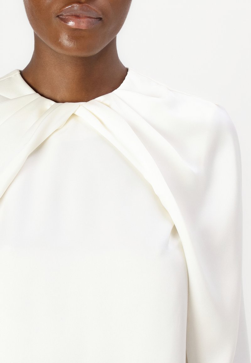Biała satynowa bluzka z węzłem przy kołnierzu, gładką fakturą i drapowanym krojem, tworząca uporządkowany, a jednocześnie swobodny wygląd.