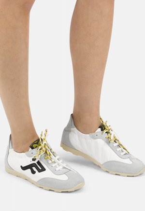 Ben iført hvide og grå sneakers med gule og sorte snørebånd, stående på en lysegrå baggrund.