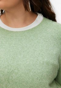 Femme portant un pull tricoté vert clair par-dessus un t-shirt blanc à col rond et une boucle d'oreille en perle pendante à l'oreille gauche.