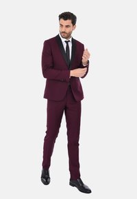 Abito borgogna con revers in satin nero, taglio elegante e pantaloni abbinati. Scarpe da sera nere completano il look formale.