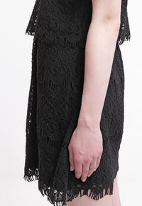 Svart spetsklänning med intrikata blommönster, fransig nederkant och figurnära design. Sidovyn visar textur och lager av tyg.