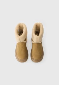 Botas de camurça castanhas com forro de lã beige, bico arredondado e sola de borracha plana. O punho dobrado destaca a suave textura interior.