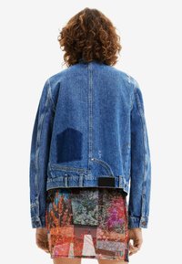 Blue denim jacket with a patch pocket and distressed details. Features a cropped cut, button closures, and is layered over a colorful patterned skirt.