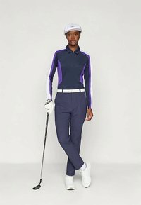 Mulher vestindo um conjunto de golfe azul-marinho e roxo, com um chapéu e sapatos brancos, segurando um taco de golfe, de pé contra um fundo neutro.