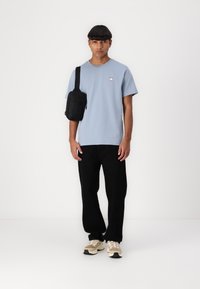 Lys blå bomulls t-skjorte med en liten rund logo, sammen med svarte løse bukser og beige sneakers. Svart crossover-bag henger på skulderen.