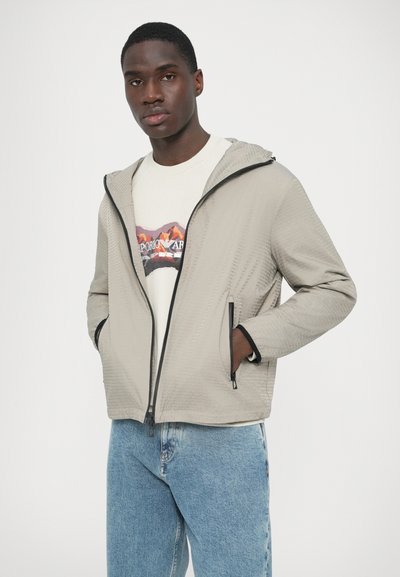 Helehall tekstuurse viimistlusega lukuga jakk, must piki tõmblukku ja varrukaid ääristav ääris, koos valge graafilise t-särgiga ja siniste teksapükstega.