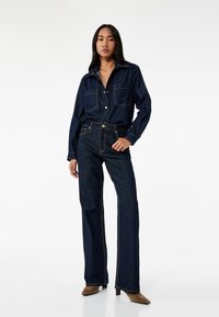 Camisa de denim con cierre de botones, dos bolsillos en el pecho y mangas largas. Pantalones vaqueros de talle alto y acampanados a juego con costuras en contraste. Botines marrones de tacón.