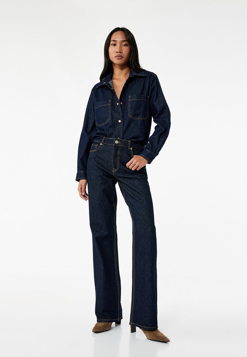 Camisa de denim con cierre de botones, dos bolsillos en el pecho y mangas largas. Pantalones vaqueros de talle alto y acampanados a juego con costuras en contraste. Botines marrones de tacón.