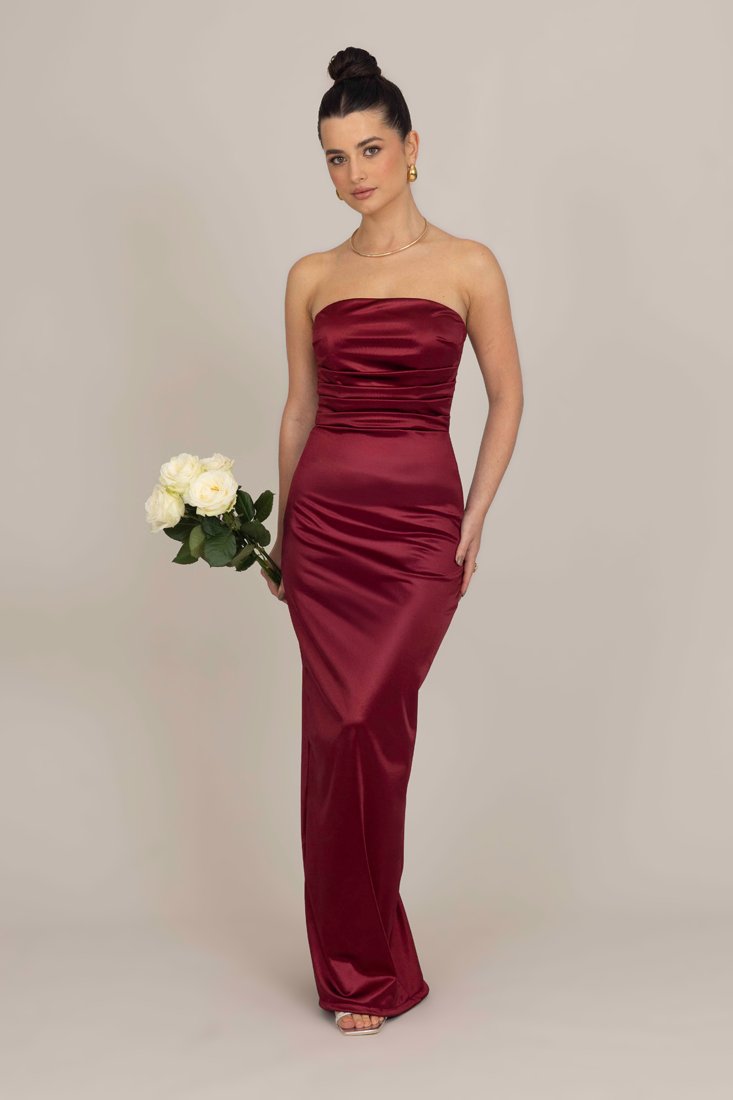 Burgunderfarbene Satin-Without-Strap-Gala mit einem gerafften Oberteil und einer taillierten Silhouette, kombiniert mit weißen Rosen und vor einem neutralen Hintergrund.