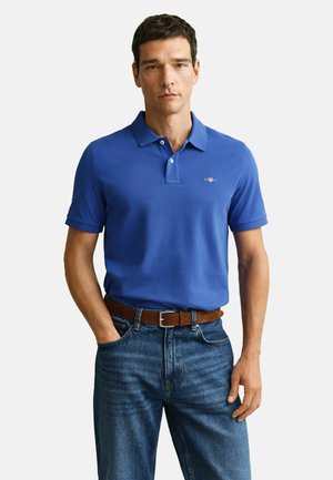 Mężczyzna w niebieskiej koszulce polo z krótkim rękawem, wpuszczonej w niebieskie dżinsy z brązowym skórzanym paskiem, stojący z jedną dłonią w kieszeni.