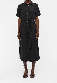 Schwarzes, knöpfbares Kleid mit kurzen Ärmeln, einem Taillengurt und seitlichen Schlitzen, aus weichem Stoff, kombiniert mit schwarzen Sandalen mit Riemen.