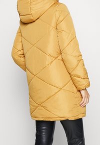 Gul puffjacka med huva, med mönster i diamantkvilt och ribbade muddar. Baksidan är slät och betonar jackans längd.