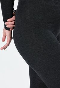 Šedé elastické legíny s měkkou texturou, které mají přiléhavý design. Ruka spočívá na oblasti boků, čímž ukazuje pohyb a přilnutí látky.