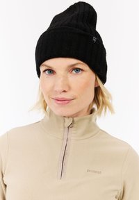 Kvinde med kort blond hår iført en sort strikhætte og en beige fleecejakke med lynlås og broderet logo på brystet.