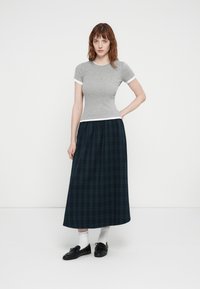Grå tettsittende t-skjorte med hvit besetning, kombinert med et langt, mørkt rutete skjørt. Modellen har på seg svarte sko og hvite sokker. Enkle, uformelle design.