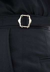 Cintura in tessuto nero con fibbia metallica argento dalla forma unica, attaccata a un capo d'abbigliamento con un sottile dettaglio testurizzato e pieghe.