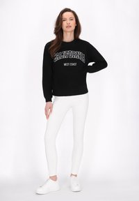 Fekete kapucnis pulóver, rajta a "CALIFORNIA" és "WEST COAST" feliratok kontrasztos fehér betűkkel, fehér skinny farmerszárral és fehér sportcipővel párosítva.