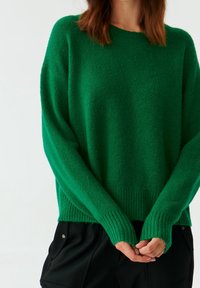 Personne portant un pull à manches longues vert en tricot et un pantalon noir, les mains jointes librement devant un fond uni.