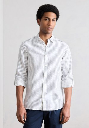 Jeune homme aux cheveux courts bouclés portant une chemise blanche à fines rayures, à manches longues retroussées, et un pantalon foncé.