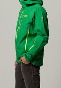 Grüne wasserdichte Jacke mit Kapuze, ausgestattet mit leuchtend gelben Reißverschlüssen, strukturiertem Finish und verstellbaren Bündchen. Sichtbare Seiten- und Brusttaschen.