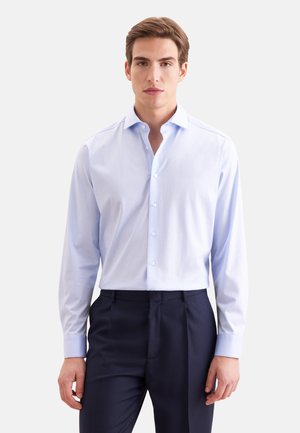 Jonge man met een lichtblauw overhemd met knopen en donkerblauwe nette broek, staand tegen een effen witte achtergrond.