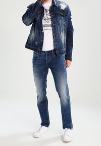 Man som bär en sliten blå denimjacka, urblekta blå jeans, en vit t-shirt med grafik och vita sneakers, står med en hand nära ansiktet.