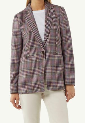 Vrouw draagt een paars-grijs geruit blazer met één knoop over een witte top en lichtbeige broek, staande met armen ontspannen langs haar zij.