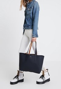 Personne portant une veste en denim, un pantalon blanc et des bottes lacées blanches, tenant un grand sac fourre-tout marine avec des poignées brunes et noires.