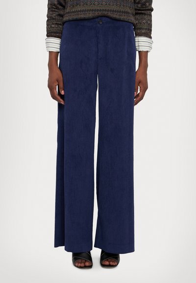 Pantalons larges en velours côtelé marine avec des côtes verticales, présentant une fermeture à bouton à l'avant et une coupe décontractée, associés à des sandales noires à lanières.