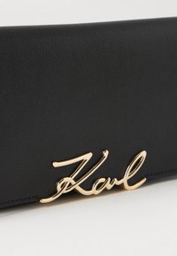 Fekete bőr pénztárca texturált felülettel és egy aranyszínű fém részlettel, amelyen a "Karl" felirat olvasható az elülső részen.