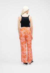 Femme blonde de dos portant un débardeur noir, un pantalon large orange à imprimé floral rose, et des baskets plateformes noires sur fond blanc.