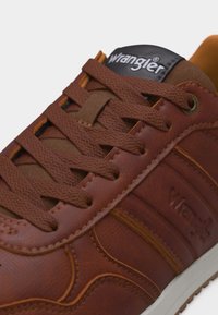 Sneaker Wrangler in pelle marrone con lacci marroni abbinati, lingua imbottita, logo in rilievo sul lato e suola bianca visibile.