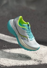 Άσπρο και τιρκουάζ παπούτσι τρεξίματος με λαμπερό κίτρινο εσωτερικό επένδυση, στον αέρα πάνω από άσφαλτο με λευκές διαβάσεις δρόμου.