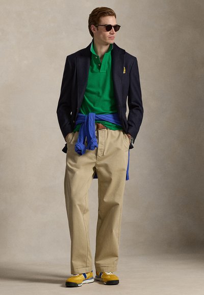 Πράσινο πόλο μπλουζάκι, μπλε σκούρο σακάκι, καφέ παντελόνι με φαρδιά μπατζάκια, μπλε πουλόβερ δεμένο στη μέση, και κίτρινα αθλητικά παπούτσια με λευκές λεπτομέρειες.