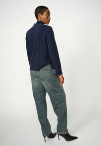 Marineblauwe gebreide trui met een hoge ribbelkraag, in combinatie met losse lichtblauwe jeans en zwarte puntige hakken. Textuurdetails van de stof zijn zichtbaar.